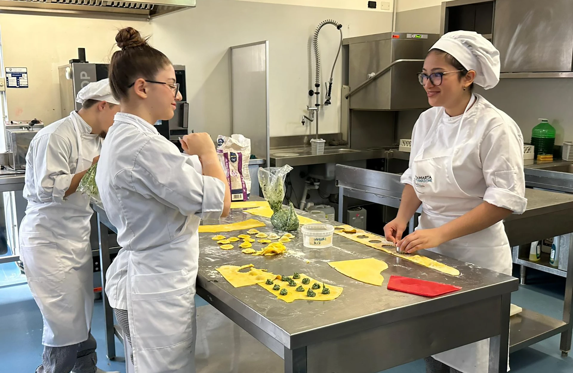 Tre studenti in divisa da chef preparano pasta fresca ripiena su un piano di lavoro in una cucina professionale durante una lezione di cucina.