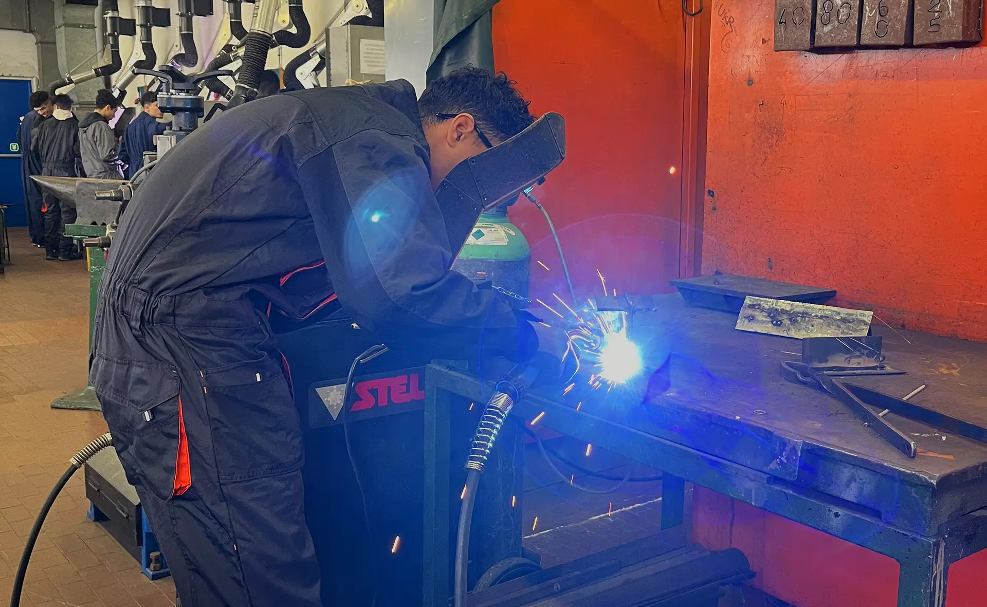 Studente in una tuta protettiva nera esegue una saldatura in un laboratorio meccanico, con scintille e luce blu provenienti dall'attività di saldatura.