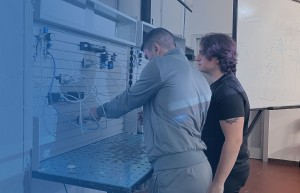 Due studenti lavorano su un pannello di controllo pneumatico in un laboratorio tecnico, con un diagramma tecnico sulla lavagna sullo sfondo.