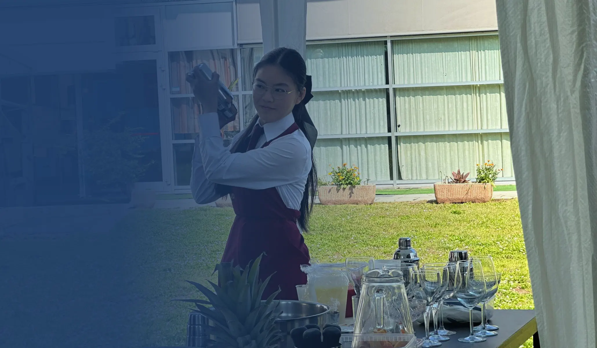 Giovane barista agita uno shaker per cocktail all'aperto, con bicchieri e strumenti da bar su un tavolo.