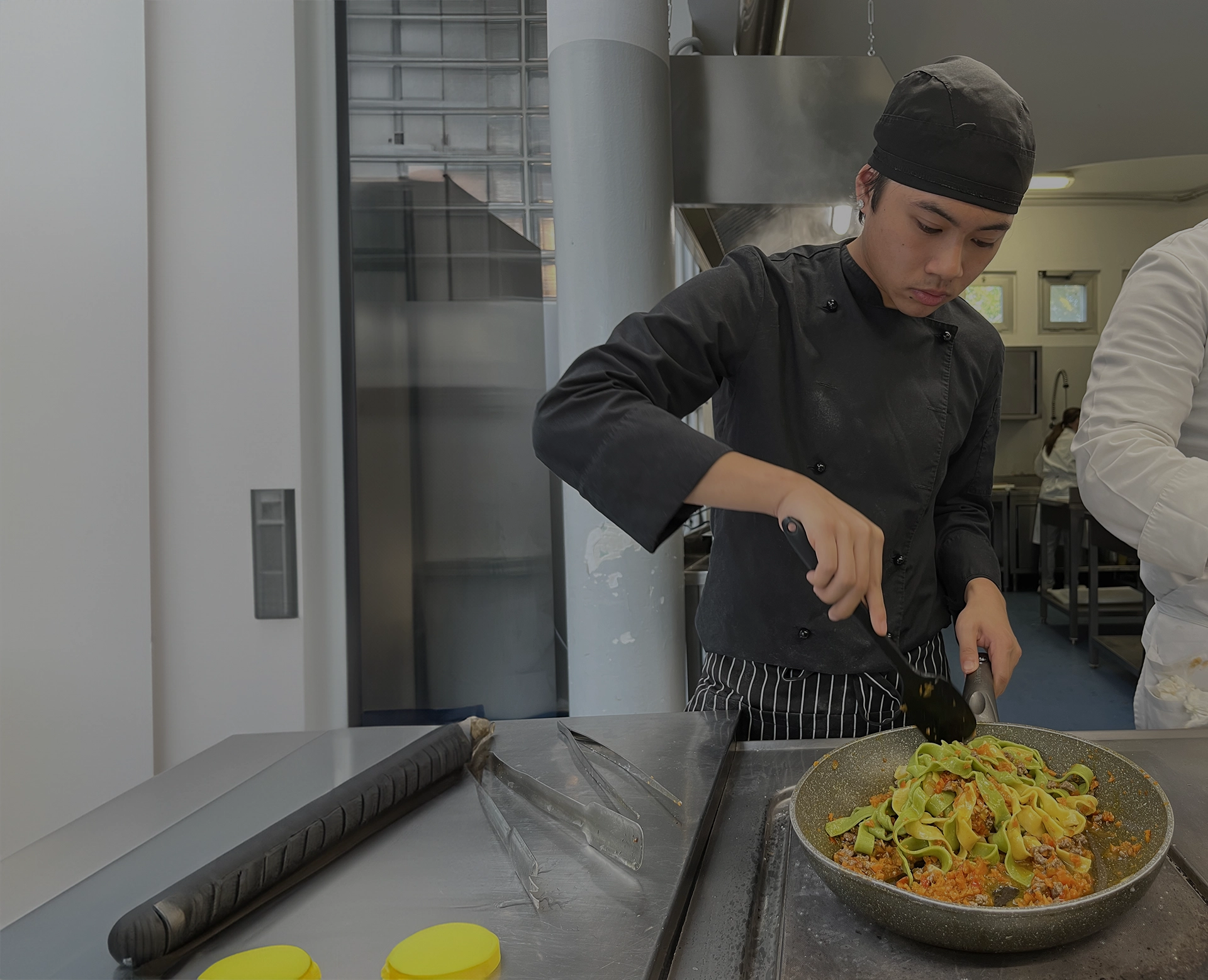 Giovane chef prepara un piatto di tagliatelle con ragù in una cucina professionale, indossando una divisa da cuoco.