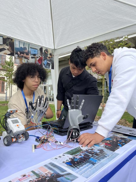 Tre studenti lavorano su un progetto di robotica, con una mano robotica e vari fili collegati a un circuito. Uno degli studenti indossa un guanto collegato al sistema per controllare la mano robotica, mentre gli altri osservano attentamente il computer.