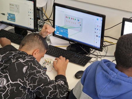 Due studenti lavorano a un progetto di elettronica utilizzando schede Arduino, con schemi di circuiti visualizzati su due monitor in laboratorio informatico.