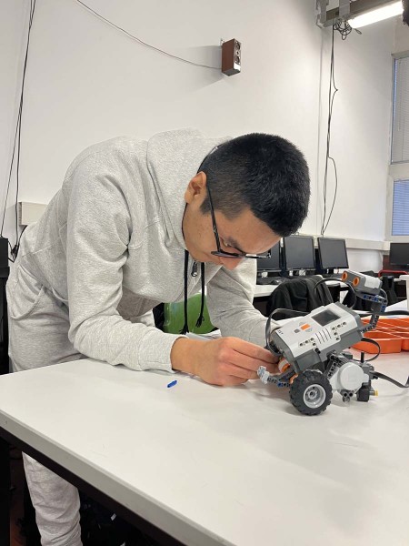 Studente concentrato sulla costruzione e programmazione di un robot LEGO Mindstorms su un tavolo in aula.