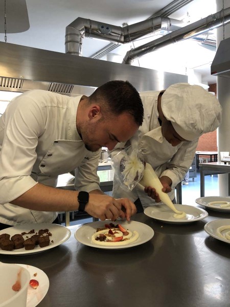 Chef e studente in cucina mentre decorano dei piatti con crema e fragole fresche, utilizzando una sac à poche per aggiungere guarnizioni artistiche su un piatto da dessert.