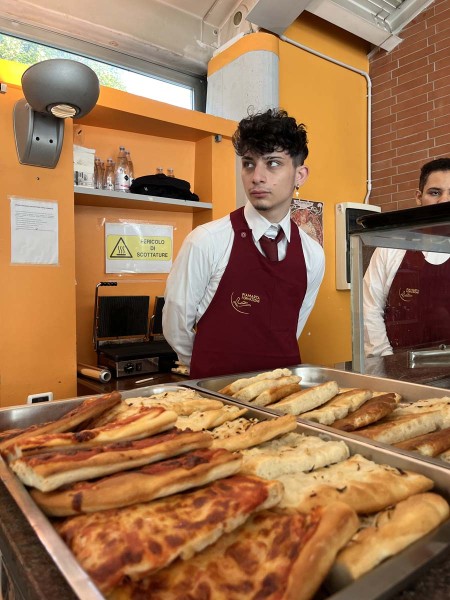 Studente in uniforme da sala, con grembiule rosso, in piedi dietro un banco di servizio con teglie di focaccia e pizza, pronto per servire i clienti in un ambiente di ristorazione.