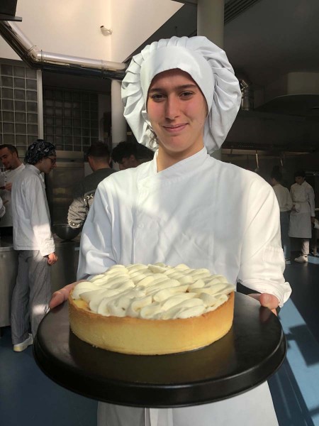 Studente di pasticceria con divisa bianca e cappello da chef, sorride mentre tiene un dolce alla crema su un vassoio nero in un ambiente di cucina scolastica.