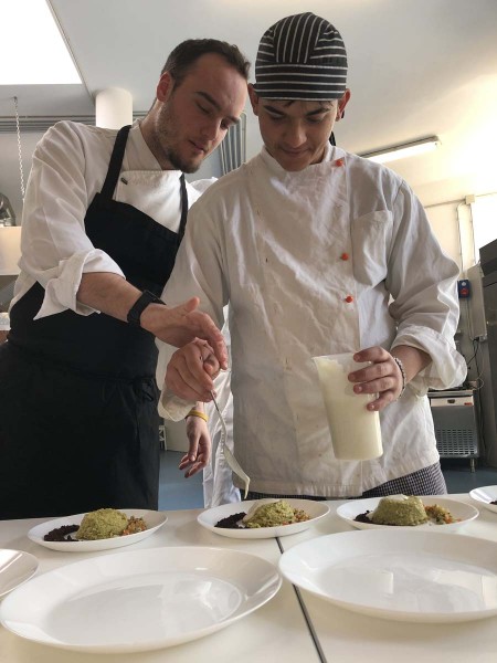 Chef guida uno studente nella preparazione di un piatto in una cucina professionale, entrambi indossano divise da cucina mentre impiattano pietanze con attenzione.