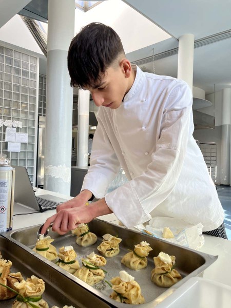 Studente in divisa da chef prepara con cura piatti gourmet disposti su una teglia in una cucina professionale.