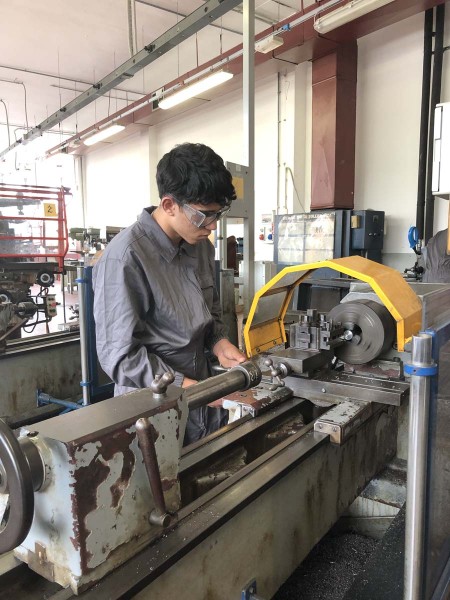 Studente con indumenti da lavoro e occhiali di protezione che utilizza un tornio meccanico in un laboratorio industriale.