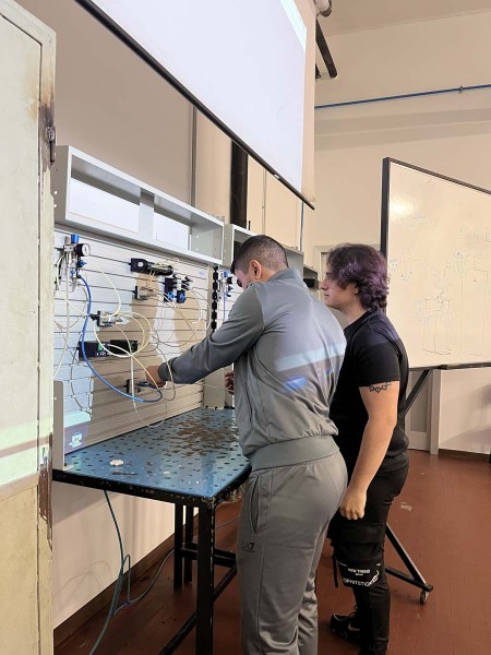 Due studenti lavorano insieme su un pannello di controllo pneumatico durante un'esercitazione pratica in laboratorio, con un'insegnante o un compagno che osserva e fornisce assistenza.