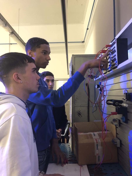 Due studenti in un laboratorio tecnico lavorano su un pannello elettrico collegando cavi durante una sessione pratica, mentre un altro studente osserva da vicino.