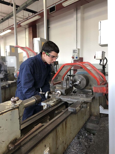 Uno studente utilizza un tornio meccanico durante un'esercitazione pratica in officina, indossando occhiali protettivi per sicurezza.