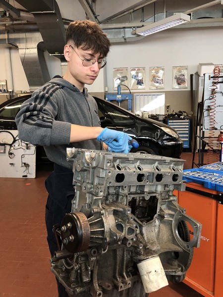 Uno studente di meccanica, con indosso guanti blu, lavora su un motore smontato all'interno di un'officina scolastica, circondato da strumenti e attrezzature meccaniche.