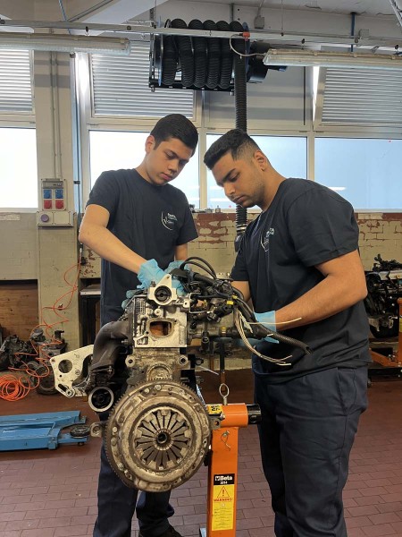 Due giovani meccanici che lavorano insieme su un motore montato su un supporto in un'officina meccanica, indossando guanti protettivi e divise da lavoro.