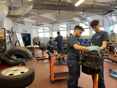 Giovani meccanici che lavorano su motori in un'officina attrezzata, indossando divise e guanti protettivi.