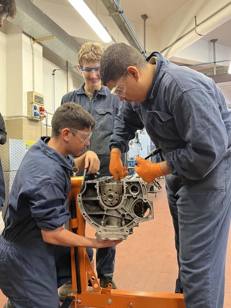Tre meccanici in tuta da lavoro e occhiali protettivi, collaborano su un componente di motore in officina.