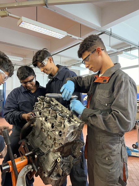 Gruppo di giovani meccanici che lavorano su un motore, indossando occhiali protettivi e guanti in officina.