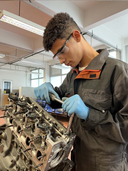 Giovane meccanico che lavora su un motore con una chiave a cricchetto, indossando guanti e occhiali protettivi.
