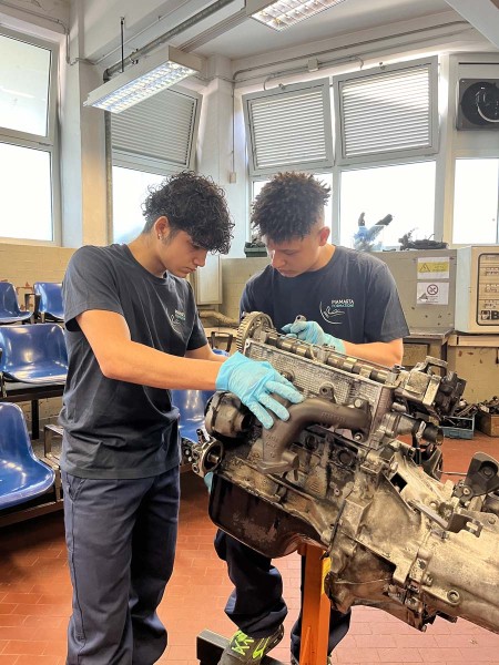 Due giovani meccanici esaminano un motore in officina, indossando guanti protettivi.