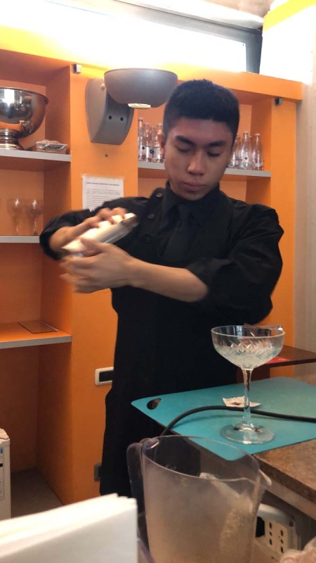 Giovane barista agita uno shaker per cocktail dietro il bancone di un bar, con un bicchiere da cocktail pronto sul bancone.