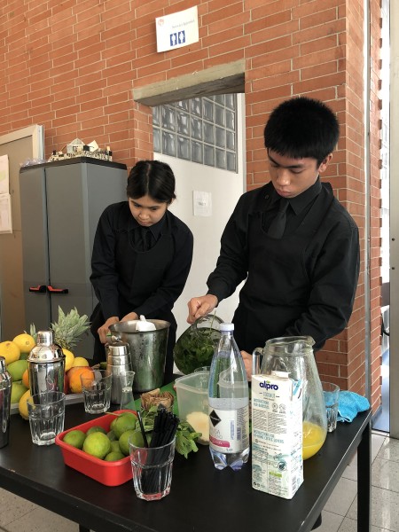 Due giovani baristi preparano bevande con ingredienti freschi su un tavolo, circondati da frutta e strumenti da bar.