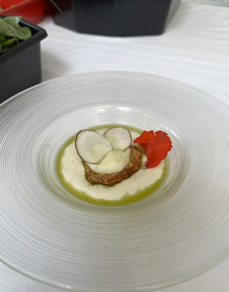 Piatto gourmet con crema bianca, verdure croccanti e guarnizione di fiori eduli su un piatto trasparente.