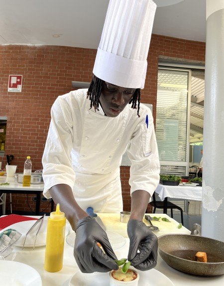 Chef in divisa bianca con guanti neri decora un piatto in una cucina professionale.