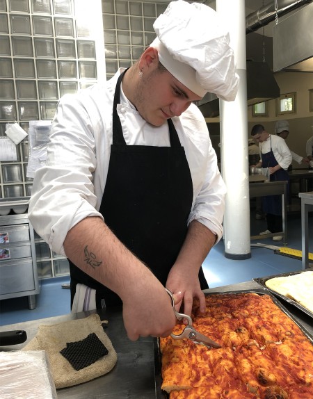 Giovane chef taglia una pizza in teglia con delle forbici in una cucina professionale.