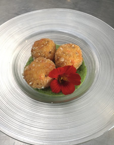 Tre polpette fritte su crema verde, guarnite con un fiore edibile rosso, servite su un piatto trasparente.