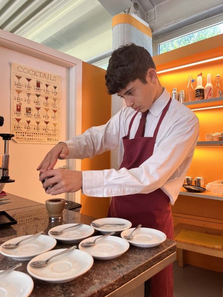 Cameriere in grembiule rosso prepara una bevanda al bancone con tazzine disposte sul tavolo