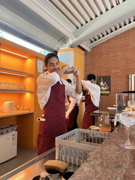 Cameriere in grembiule rosso agita uno shaker dietro il bancone insieme ad altri camerieri