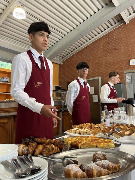 Camerieri in grembiule rosso in piedi vicino a un buffet con croissant, pane e bevande