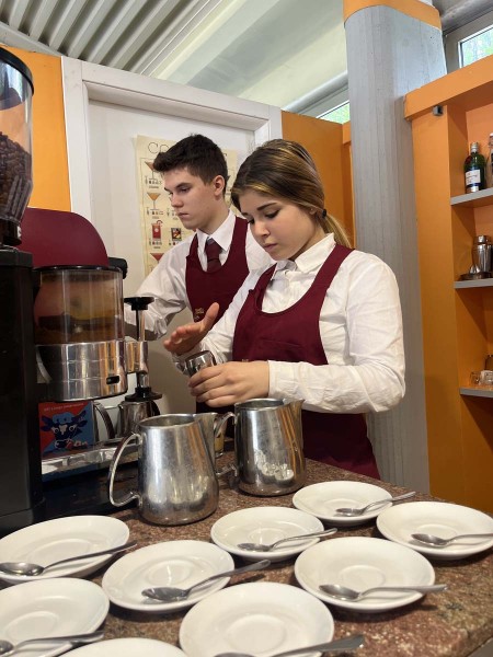 Due camerieri in grembiule rosso preparano caffè dietro il bancone con tazzine e cucchiaini pronti sul tavolo