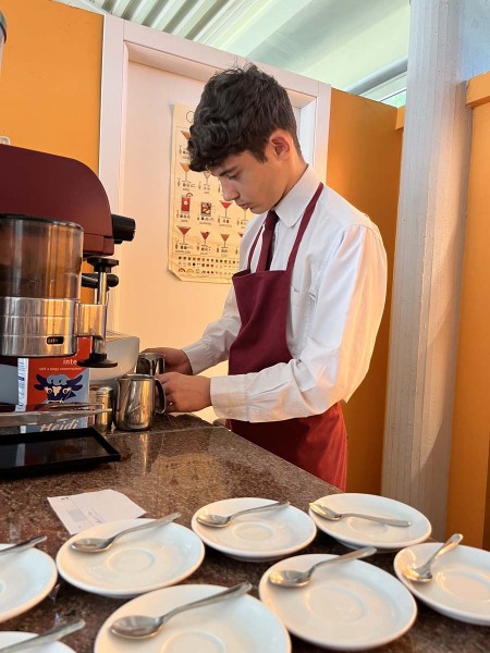 Cameriere in grembiule rosso prepara il caffè con una macchina da espresso, tazzine e cucchiaini pronti sul tavolo