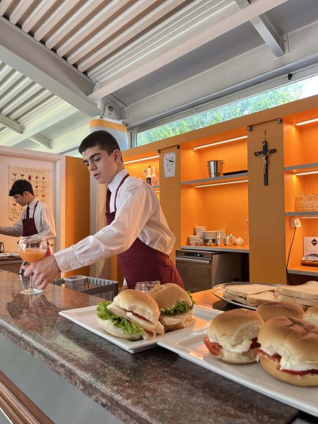 Cameriere in grembiule rosso prepara un bicchiere di cocktail al bancone, con panini disposti su un piatto